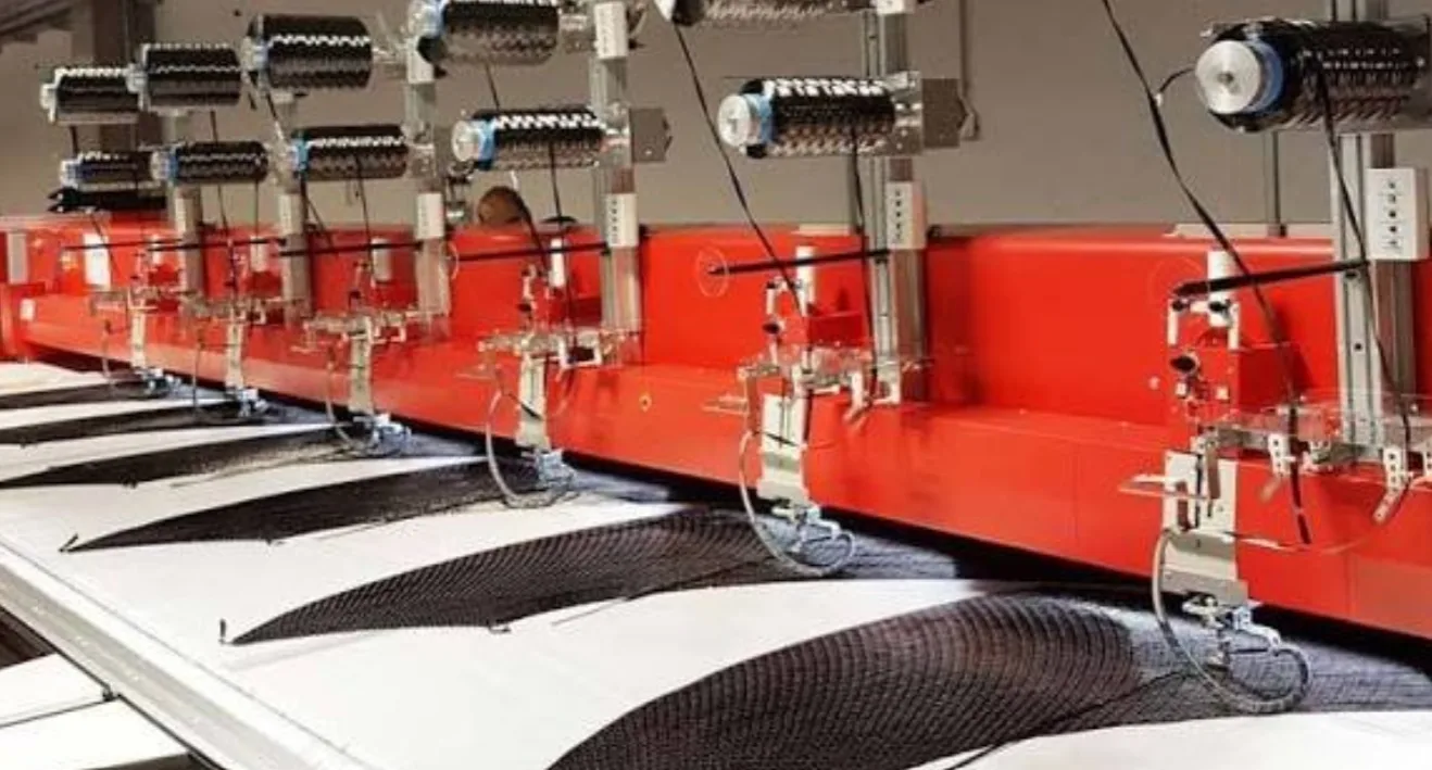 A row of multi-head tailored fiber placement machines stitching black carbon fiber tow into curved blade-shaped preforms on white backing fabric.