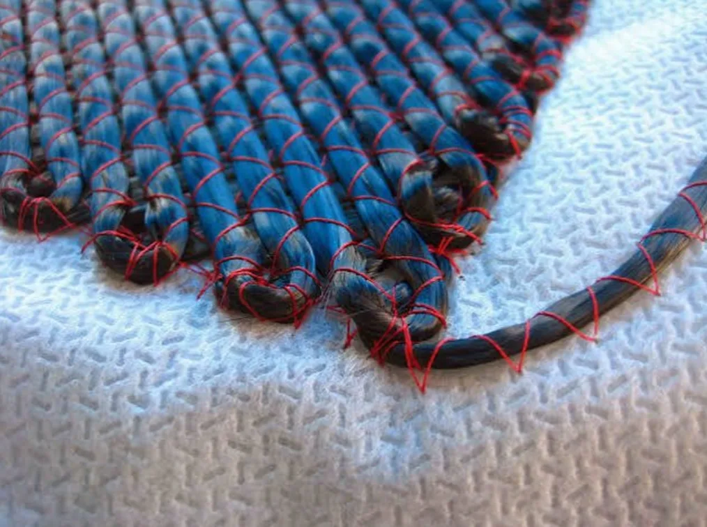 Close-up of carbon fiber tow couched onto a white backing cloth with red stitching thread, showing the tight loops that hold each fiber path in place.