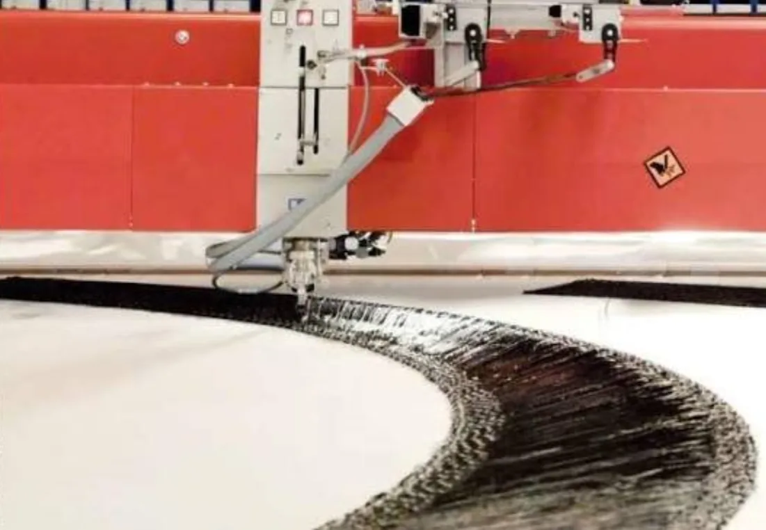 A single stitching head laying a continuous curved path of carbon fiber tow onto white backing material.